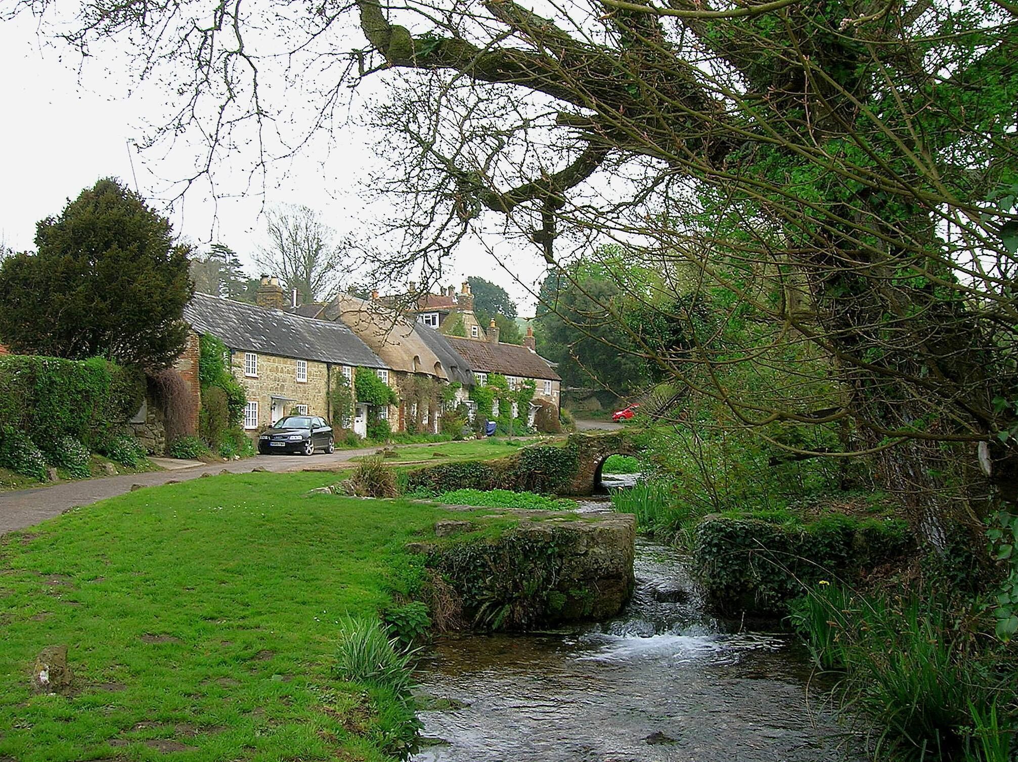 A rural village with stone houses, a narrow road, and a stream with a bridge.