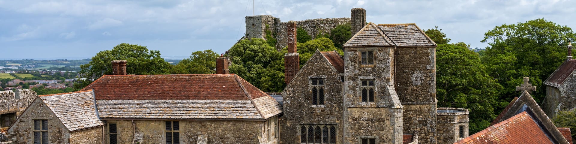 A historic stone building with a red-tiled roof, surrounded by lush greenery and a cloudy sky.