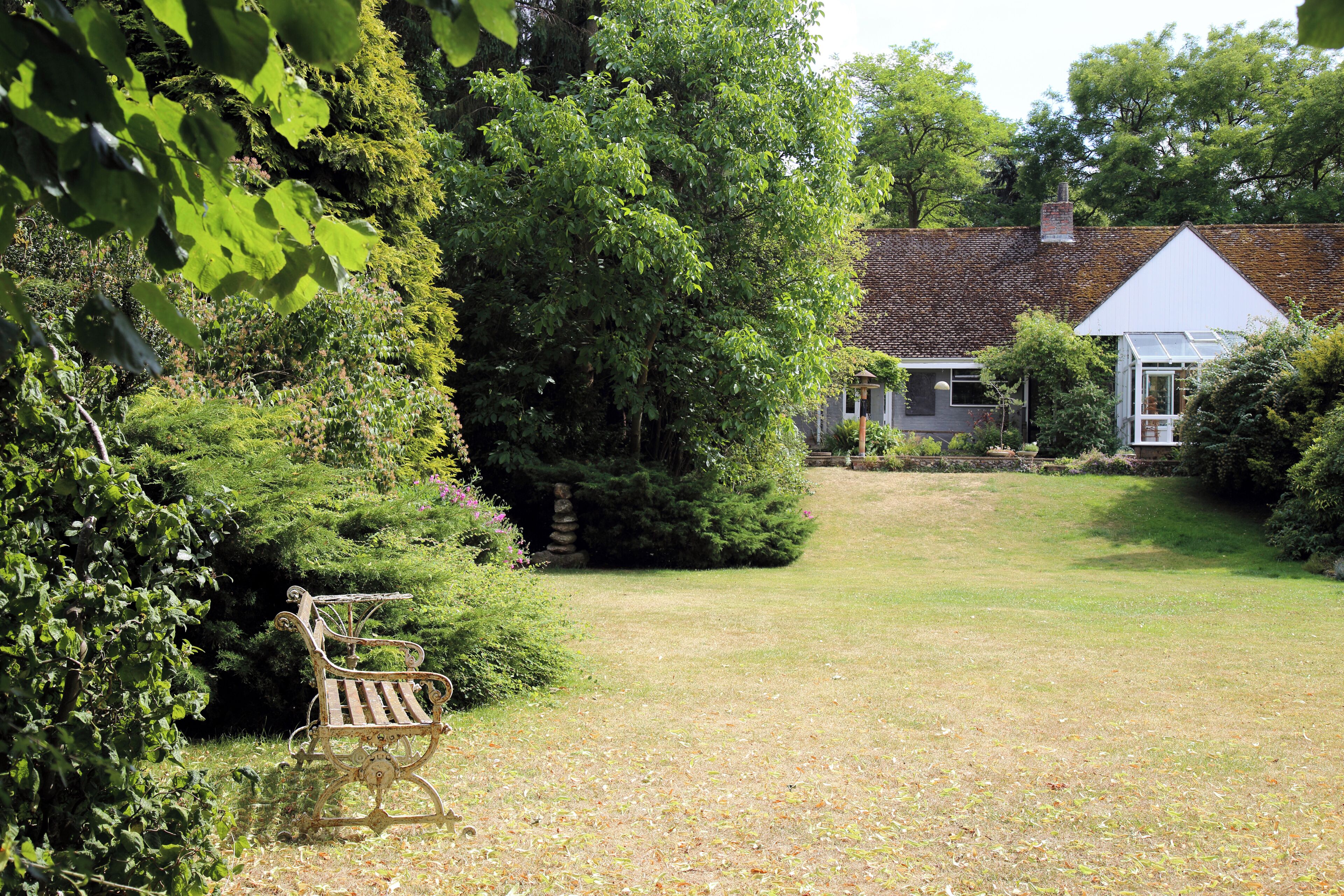 A lawn and garden bench backed by shrubs, trees and tiled-roof building at The Gibberd Garden, in the Harlow District of Essex, England