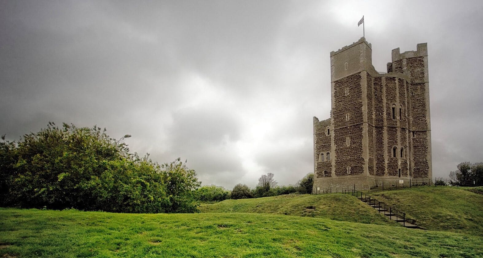 A historic stone tower with a flag, surrounded by lush green grass and trees under a cloudy sky.