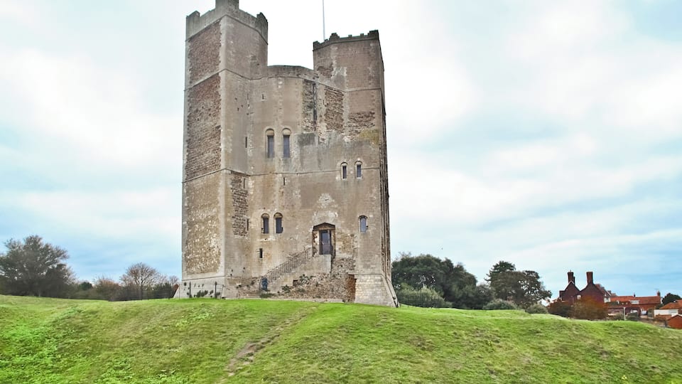 A historic stone tower with battlements, set against a grassy hill and a partly cloudy sky.