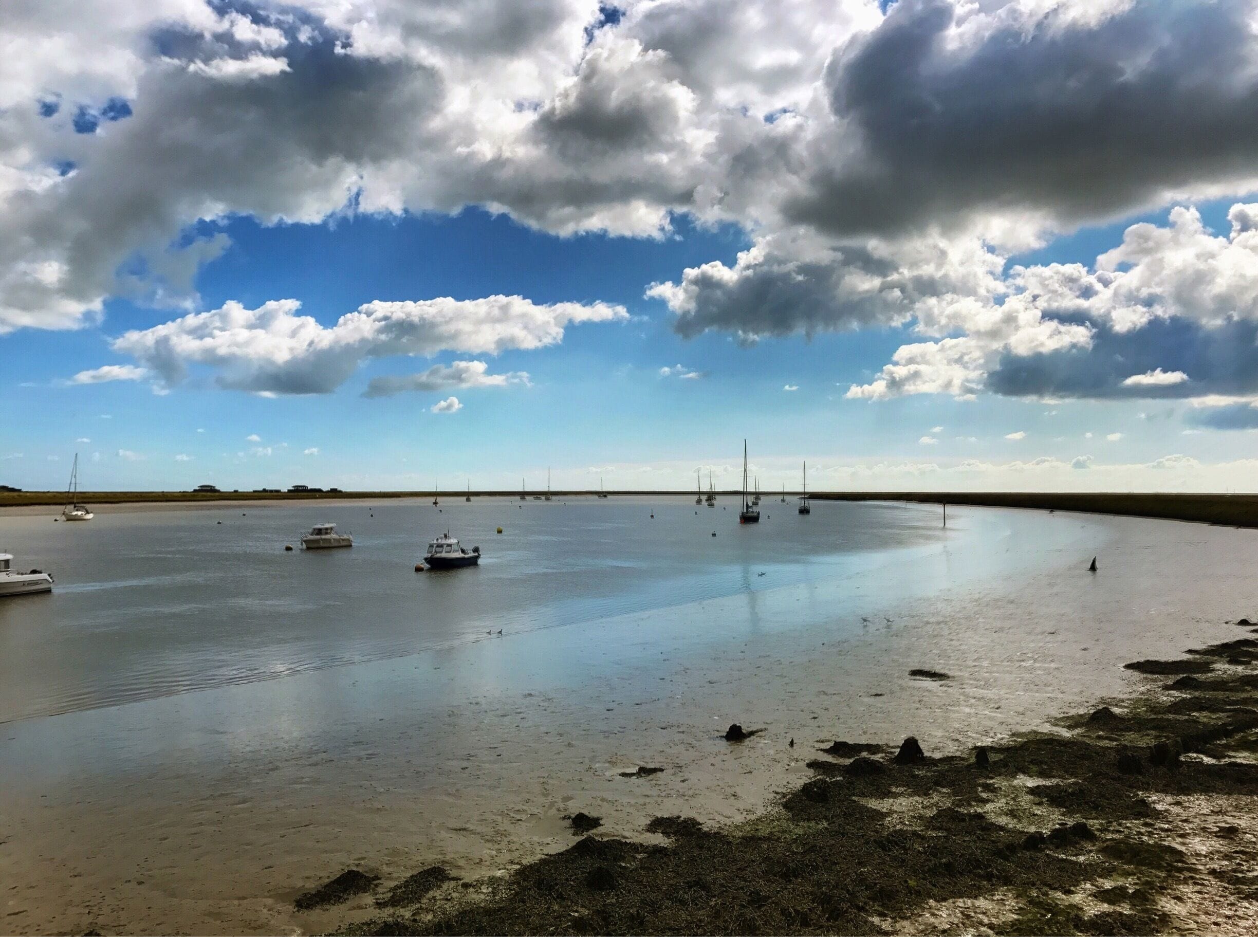 A calm bay with several boats anchored, a sandy shore with seaweed, and a partly cloudy sky.