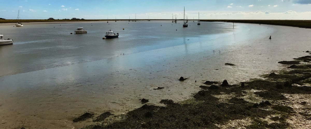 A calm bay with several boats anchored, a sandy shore with seaweed, and a partly cloudy sky.