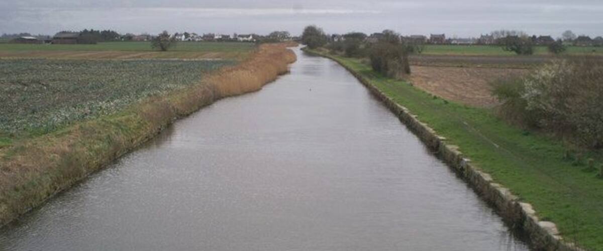 A narrow waterway bordered by fields and a cloudy sky.