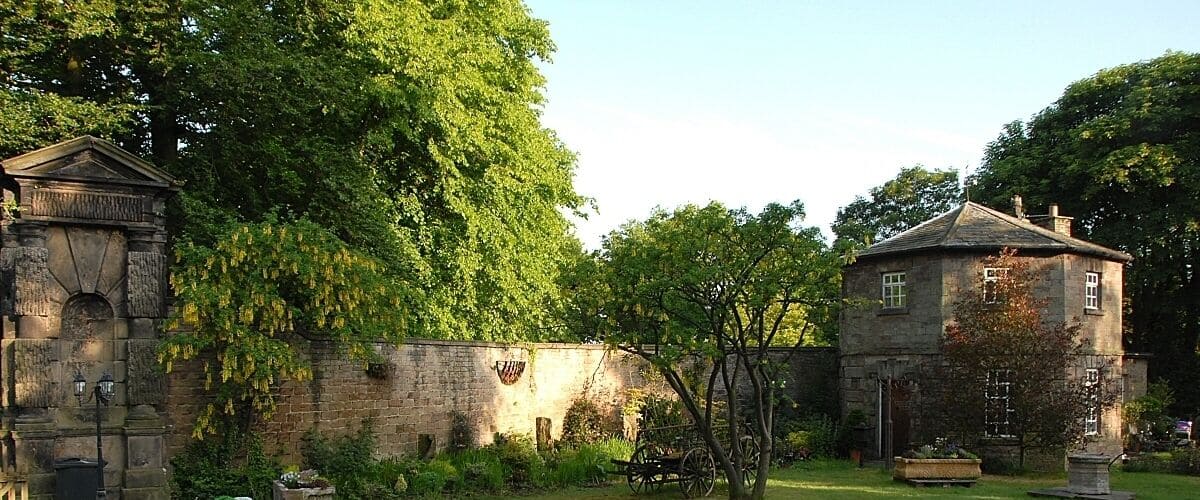 A historic stone building with a garden and a large tree in the background.