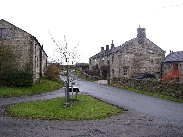 A narrow rural road flanked by stone buildings, a grassy area, and a parked car.