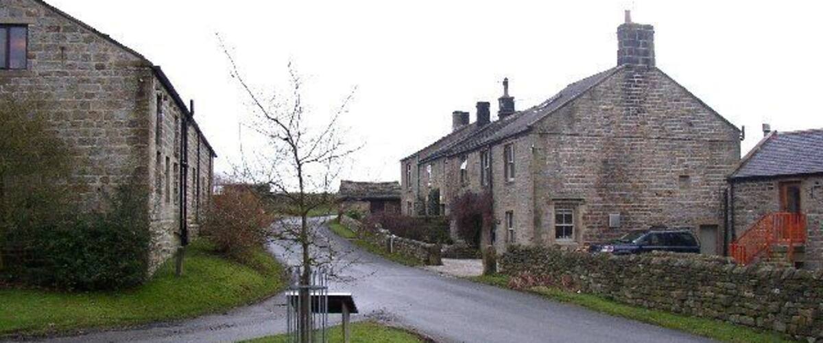 A narrow rural road flanked by stone buildings, a grassy area, and a parked car.