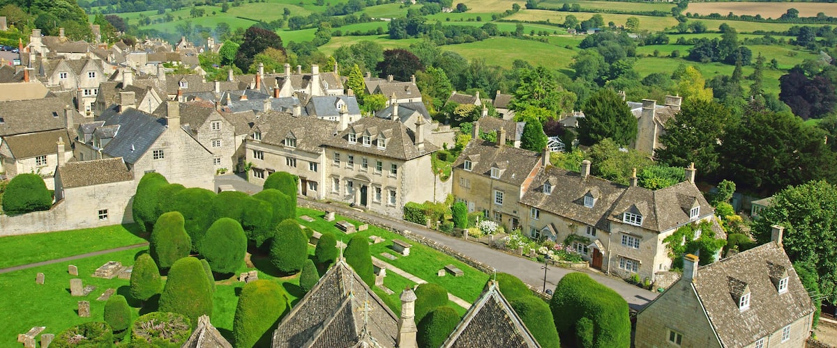 A historic village with stone buildings, a well-maintained garden, and rolling green hills in the background.