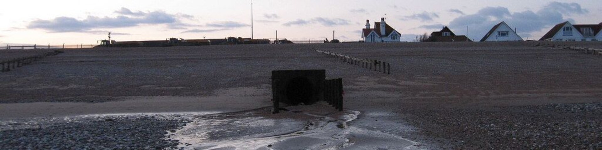 Una playa con un pequeño estanque, una cerca de madera y casas al fondo.