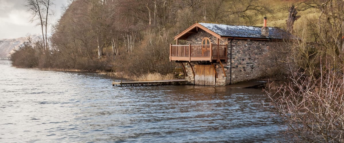 A wooden boathouse by a river, surrounded by trees and hills.