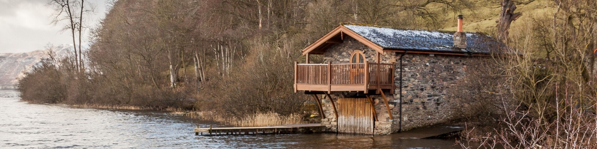 A wooden boathouse by a river, surrounded by trees and hills.