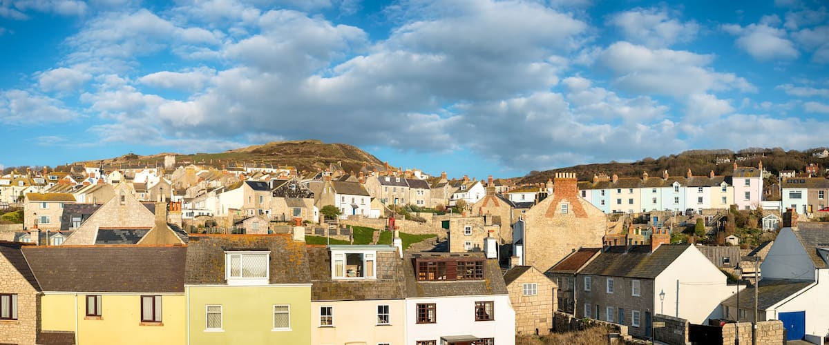 A coastal town with colorful houses, a prominent hill in the background, and a clear blue sky.
