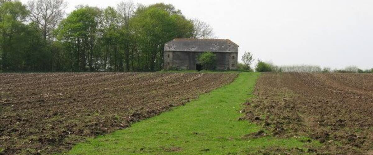 A rural landscape with a plowed field, a grassy path, and a stone building in the distance.