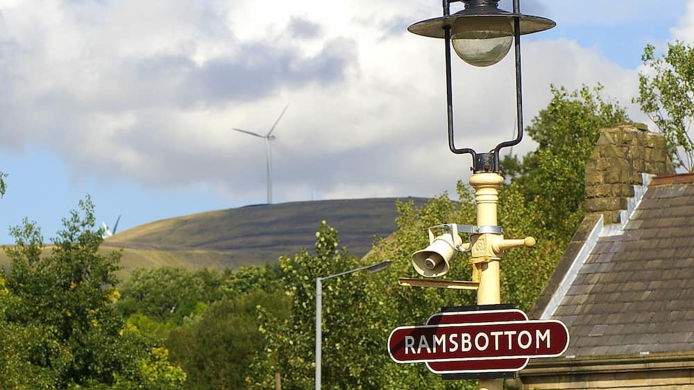 A street lamp with a 'Ramsbottom' sign, a wind turbine, and lush greenery in the background.