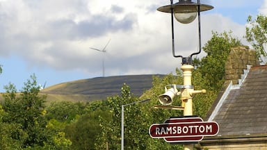Um poste de luz com uma placa 'Ramsbottom', uma turbina e vegetação exuberante ao fundo.
