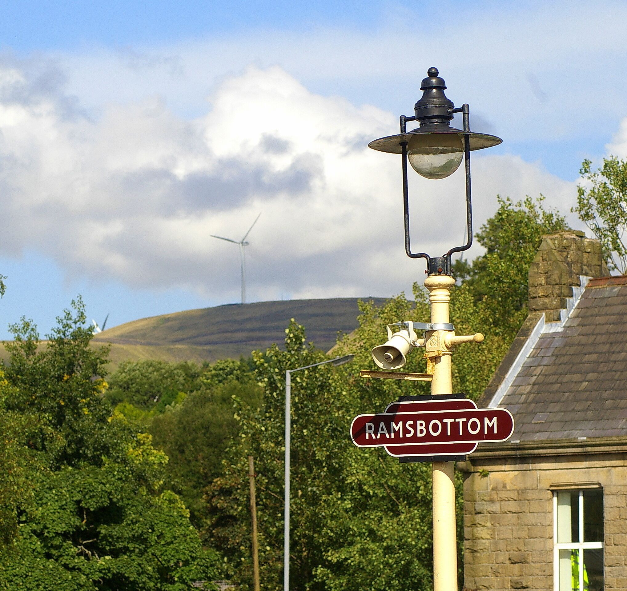 A street lamp with a 'Ramsbottom' sign, a wind turbine, and lush greenery in the background.