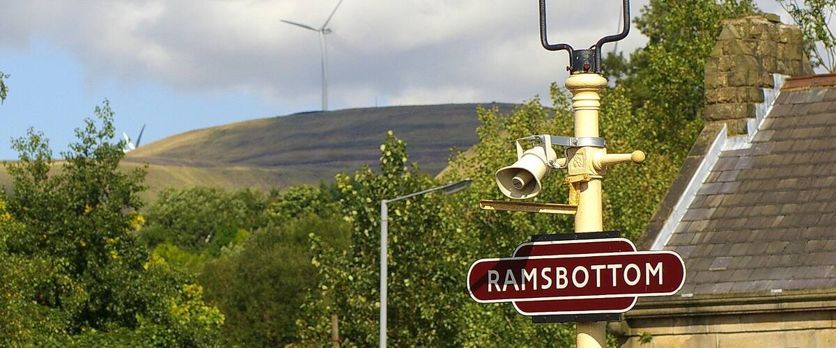 A street lamp with a 'Ramsbottom' sign, a wind turbine, and lush greenery in the background.
