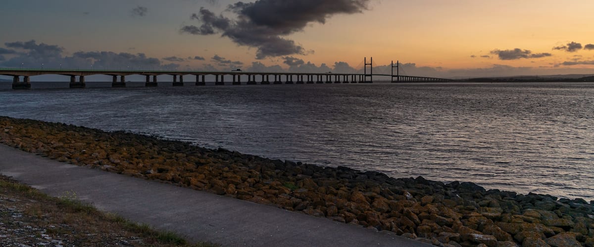 A bridge over water with a rocky shoreline and a cloudy sky.