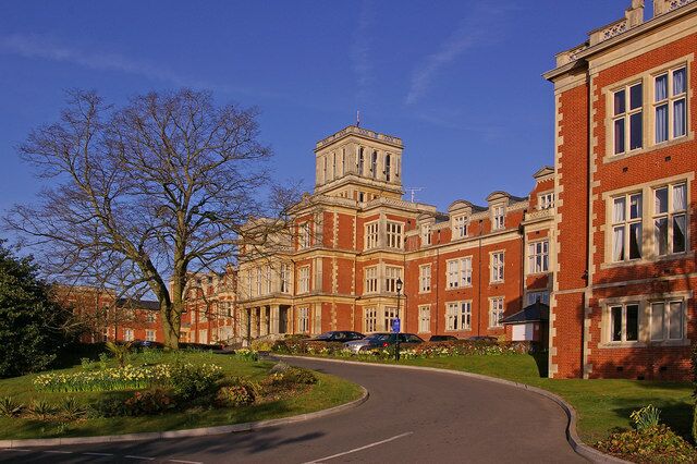 A large red-brick building with a tower, surrounded by a curved road and a tree.