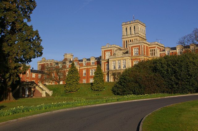 A large, historic building with a prominent tower, surrounded by lush greenery and a curved road.