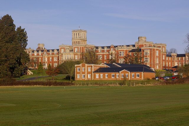 A large red brick building with a prominent tower, surrounded by green fields and trees.