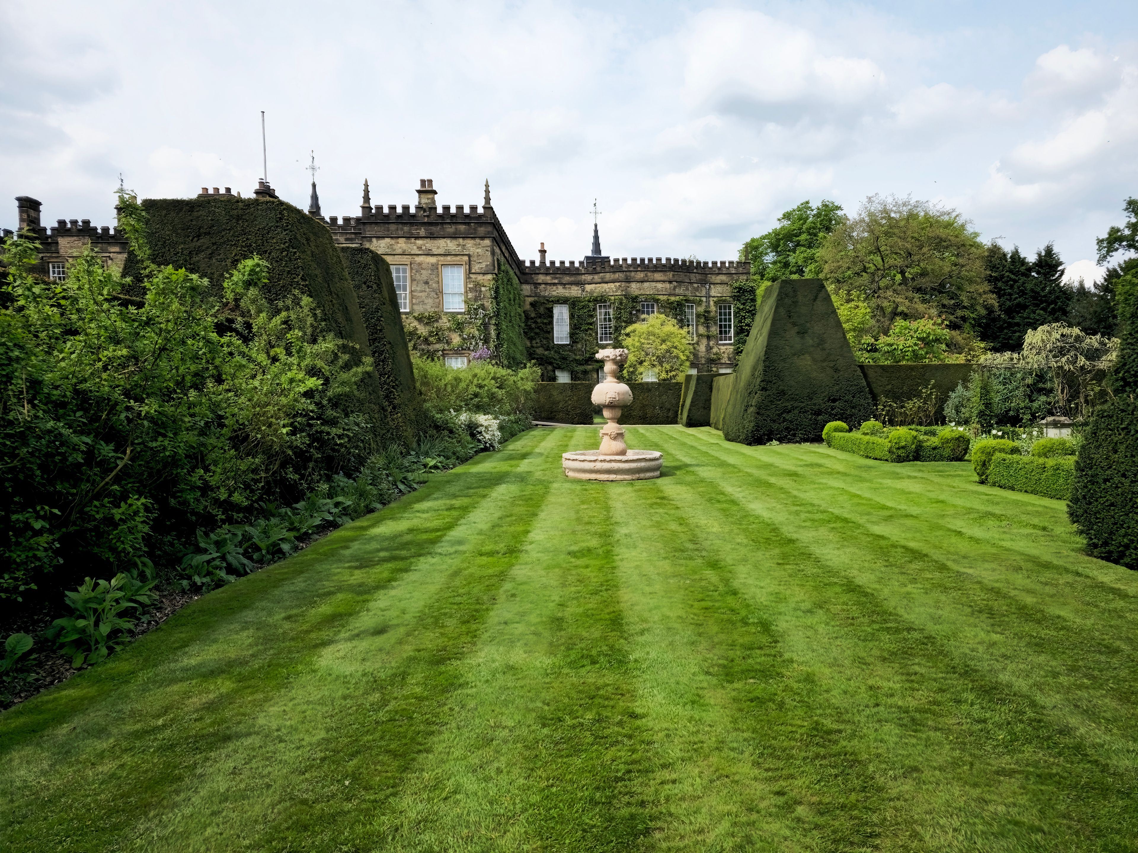 A well-manicured lawn with a central fountain, surrounded by neatly trimmed hedges and a historic building with a turret.