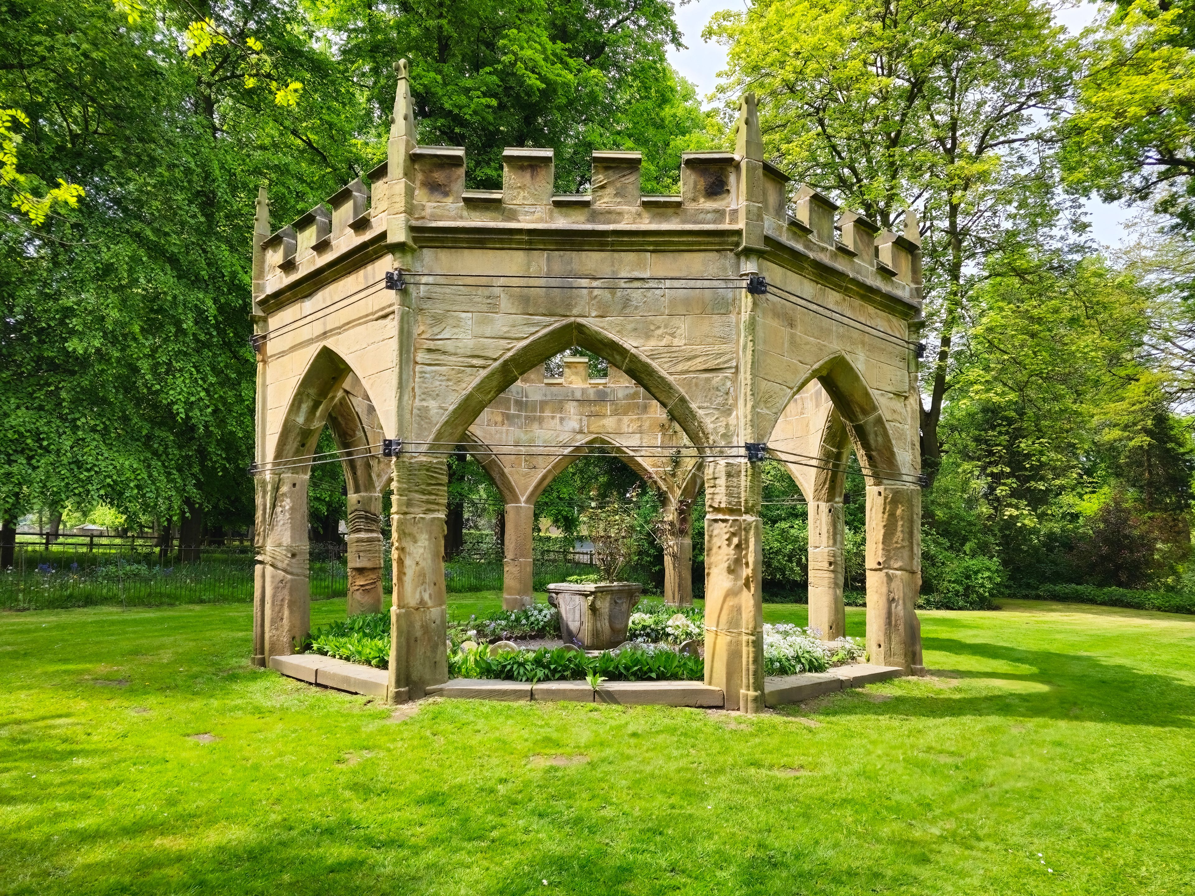 A stone gazebo with pointed arches and a crenellated roof, surrounded by lush greenery and a well-maintained lawn.