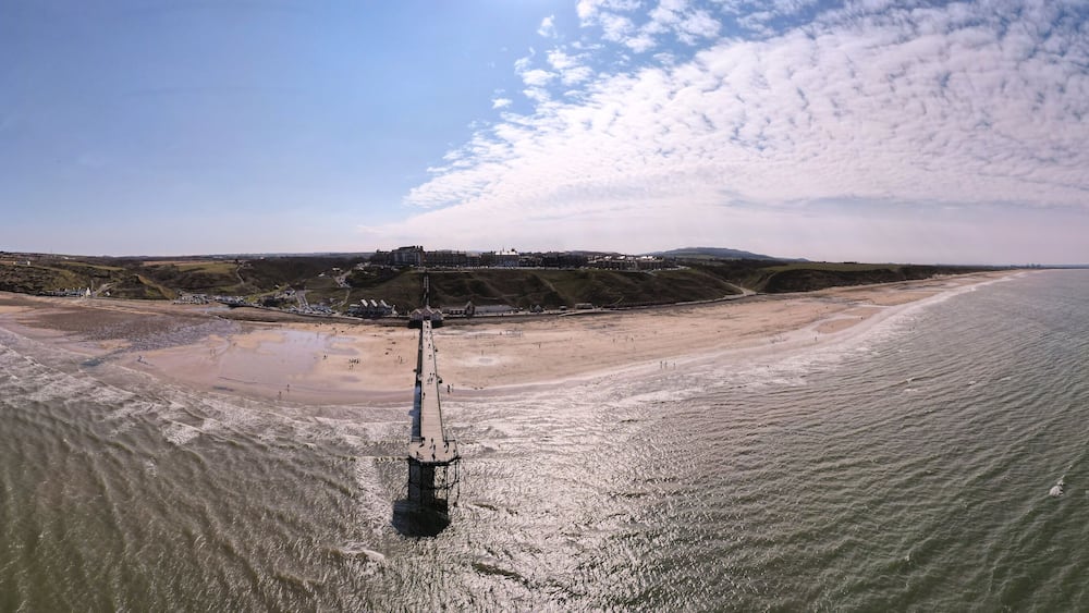 An aerial view of a beach with a pier extending into the water, a sandy shore, and a distant hill with buildings.