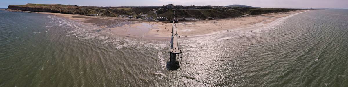 An aerial view of a beach with a pier extending into the water, a sandy shore, and a distant hill with buildings.