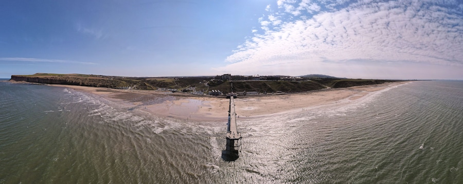 An aerial view of a beach with a pier extending into the water, a sandy shore, and a distant hill with buildings.
