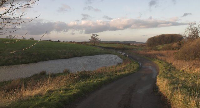A rural landscape with a narrow road curving alongside a river, surrounded by fields and trees.