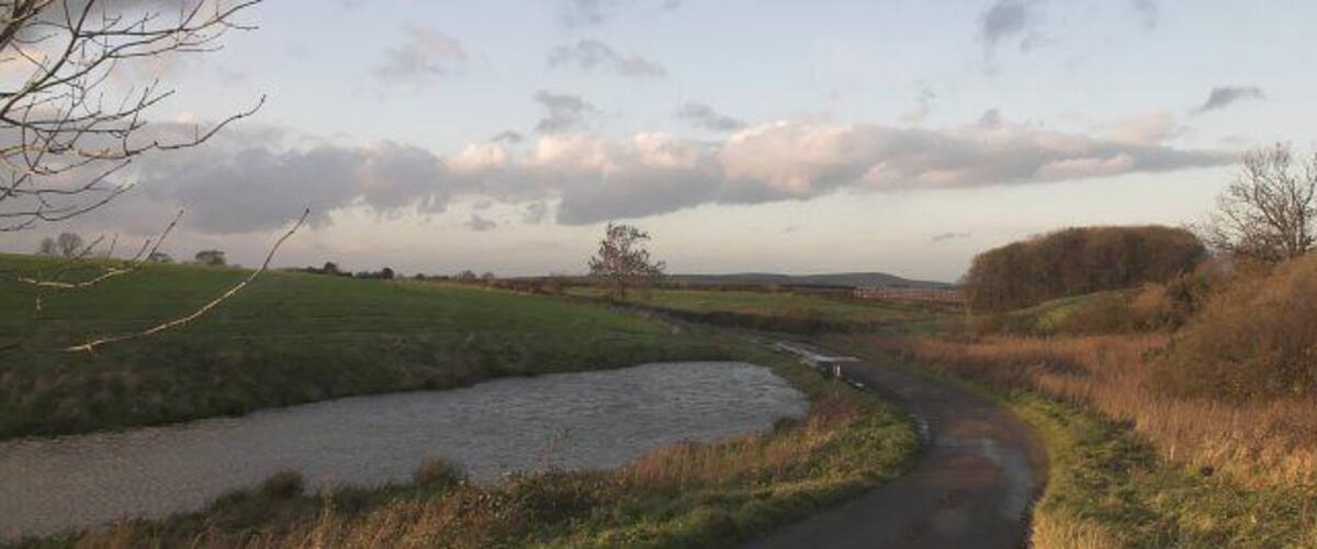 A rural landscape with a narrow road curving alongside a river, surrounded by fields and trees.