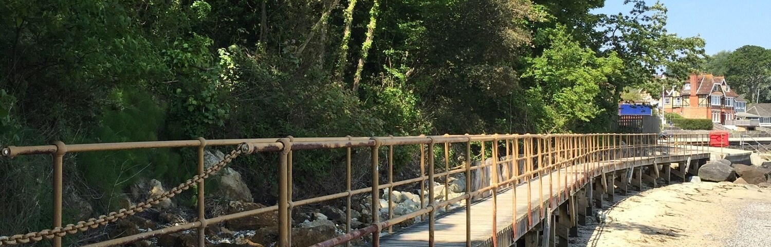 A wooden bridge over a rocky beach with trees on one side and houses on the other.