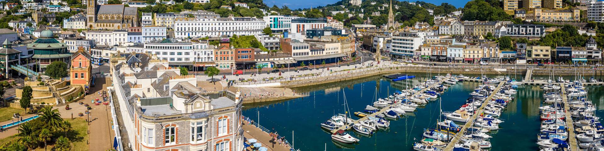 Aerial view of Torquay Marina on a sunny day, featuring yachts, boats, promenade, and blue sea along the English Riviera coastline