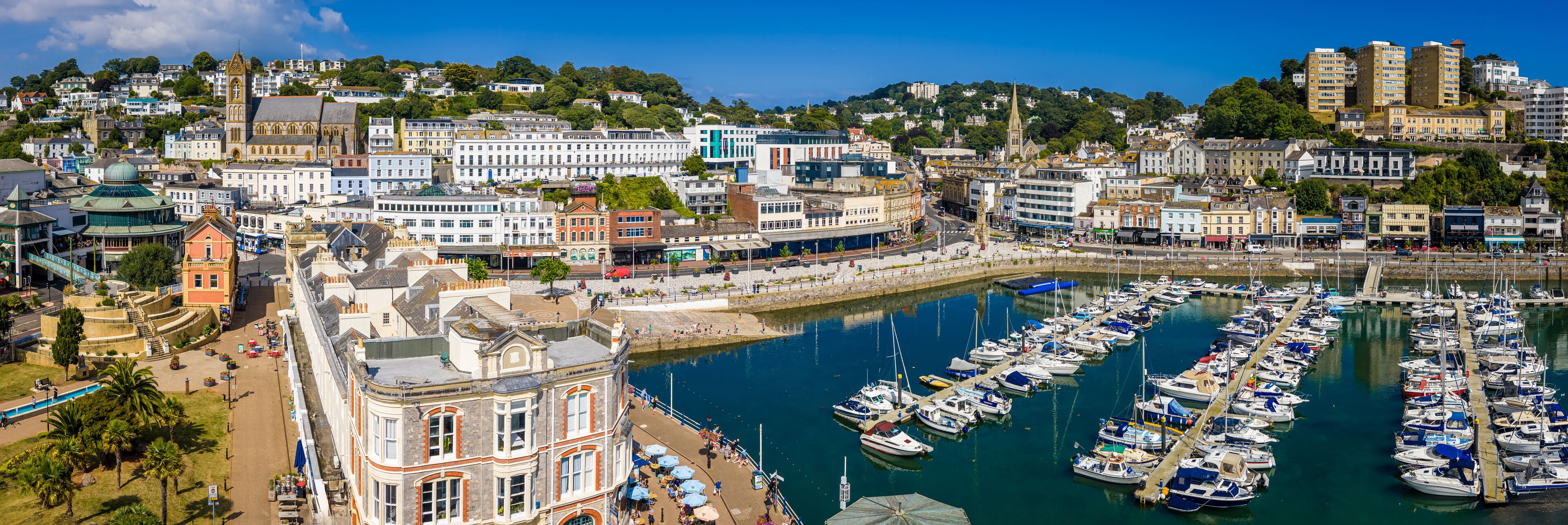 A harbor with numerous boats docked, a promenade with people, and historic buildings along the waterfront.