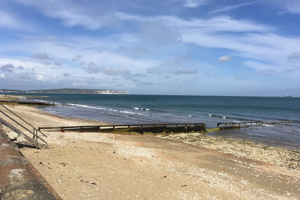Une plage avec une promenade en bois, des galets et une falaise qui se dessine à l’horizon.