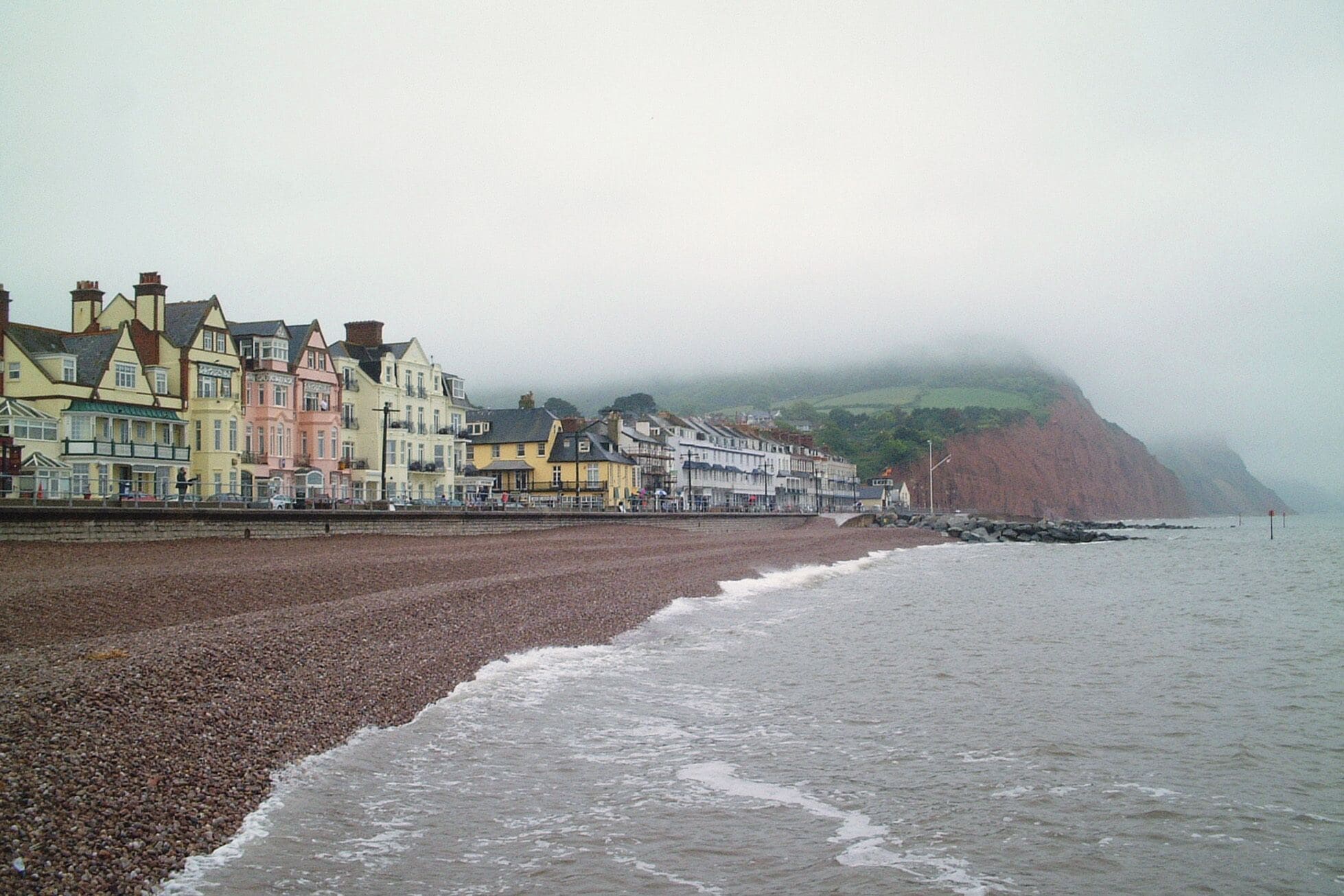 A pebble beach with colorful buildings and a cliff in the background.