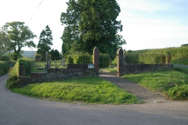 A gated rural road with a stone wall and metal gate, surrounded by greenery and trees.