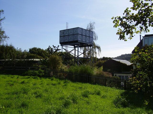 A water tower in a grassy field with a fence and trees in the background.