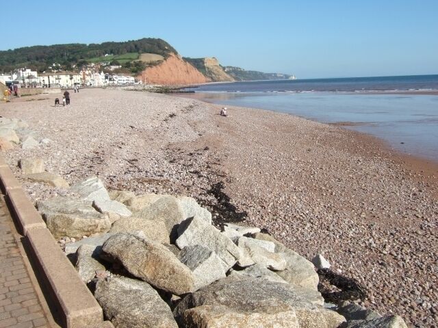 A pebbly beach with large rocks in the foreground, a distant cliff, and a clear sky.