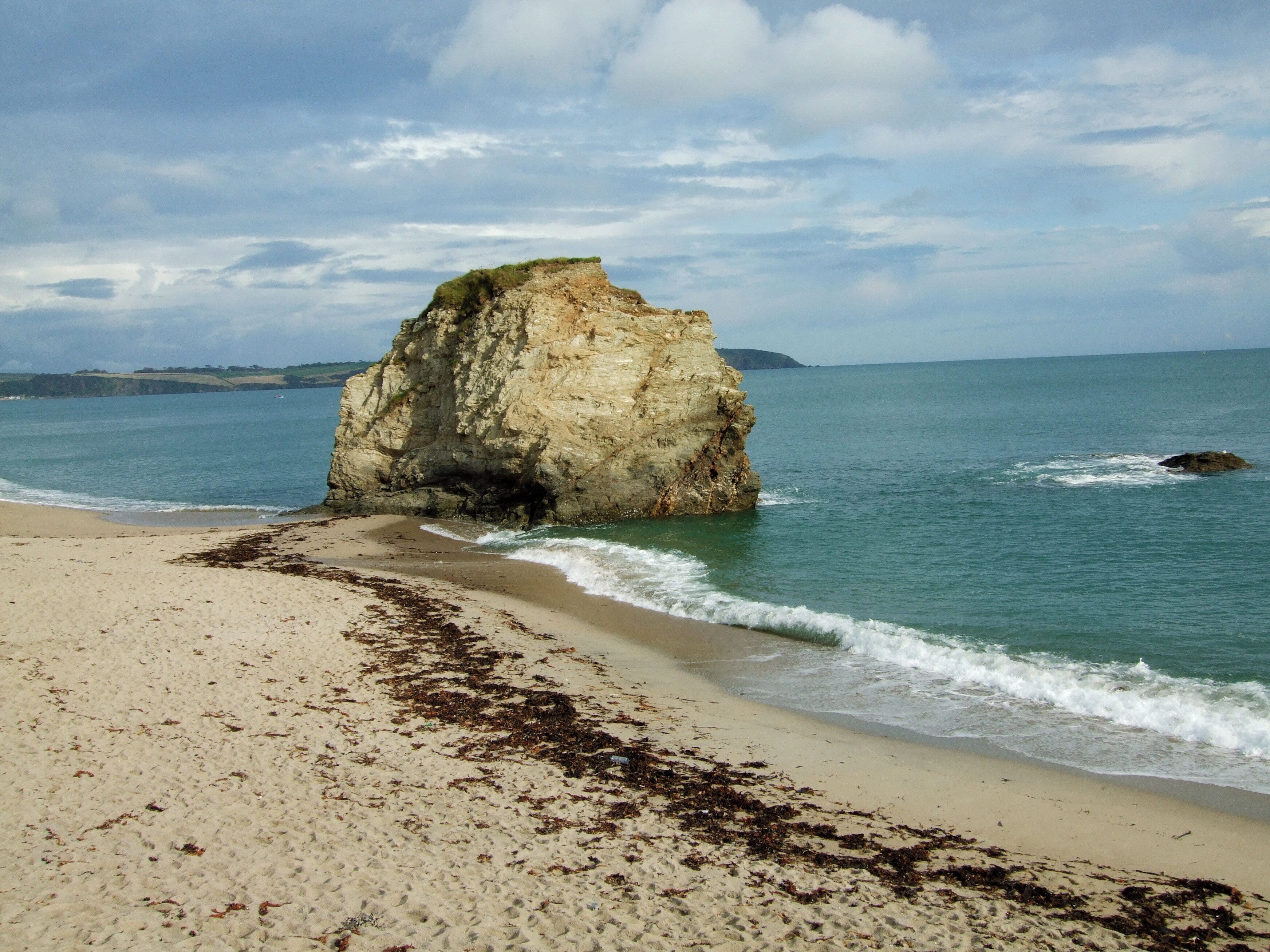 A sandy beach with seaweed, a large rock formation, and calm ocean waves.