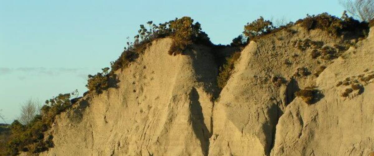 A large, steep cliff with sparse vegetation at the top and a fenced area at the base.