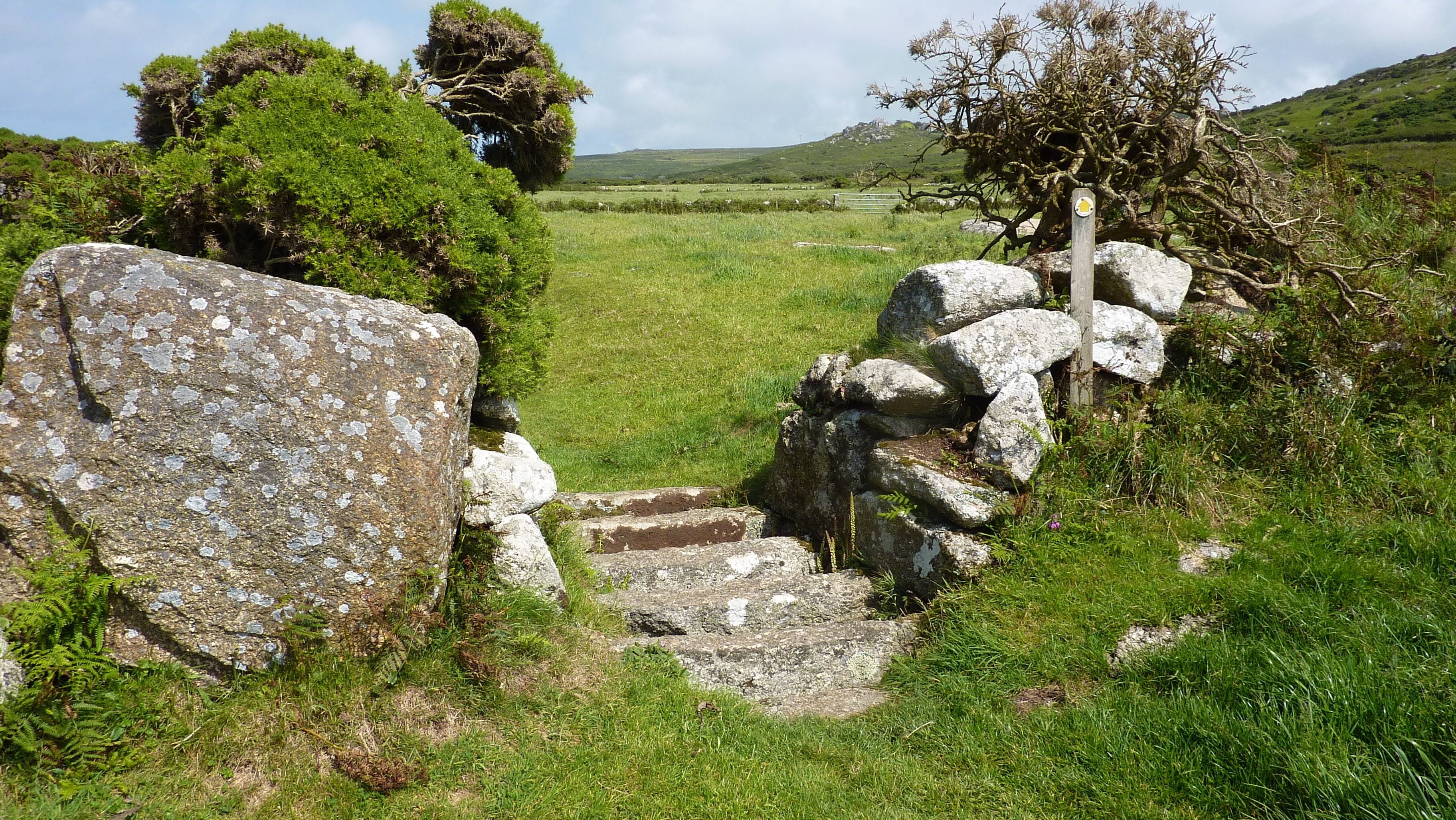 A stone gate in a grassy field with trees and hills in the background.