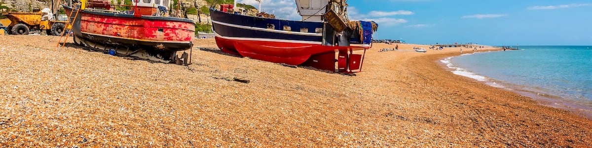 A pebbled beach with fishing boats, a lighthouse, and a coastal building.