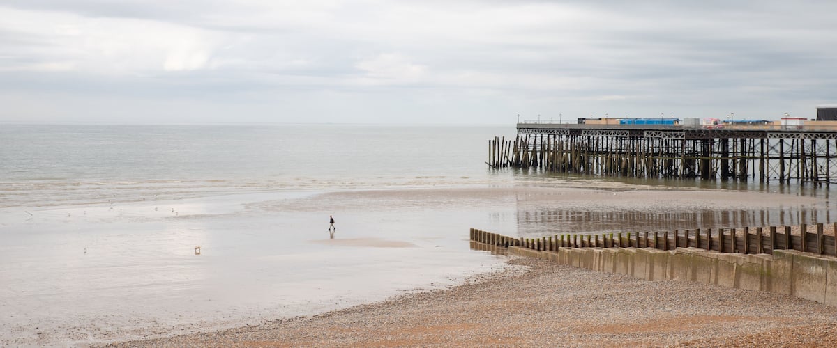 A beach with a wooden pier extending into the water, a person walking along the shore, and a cloudy sky.