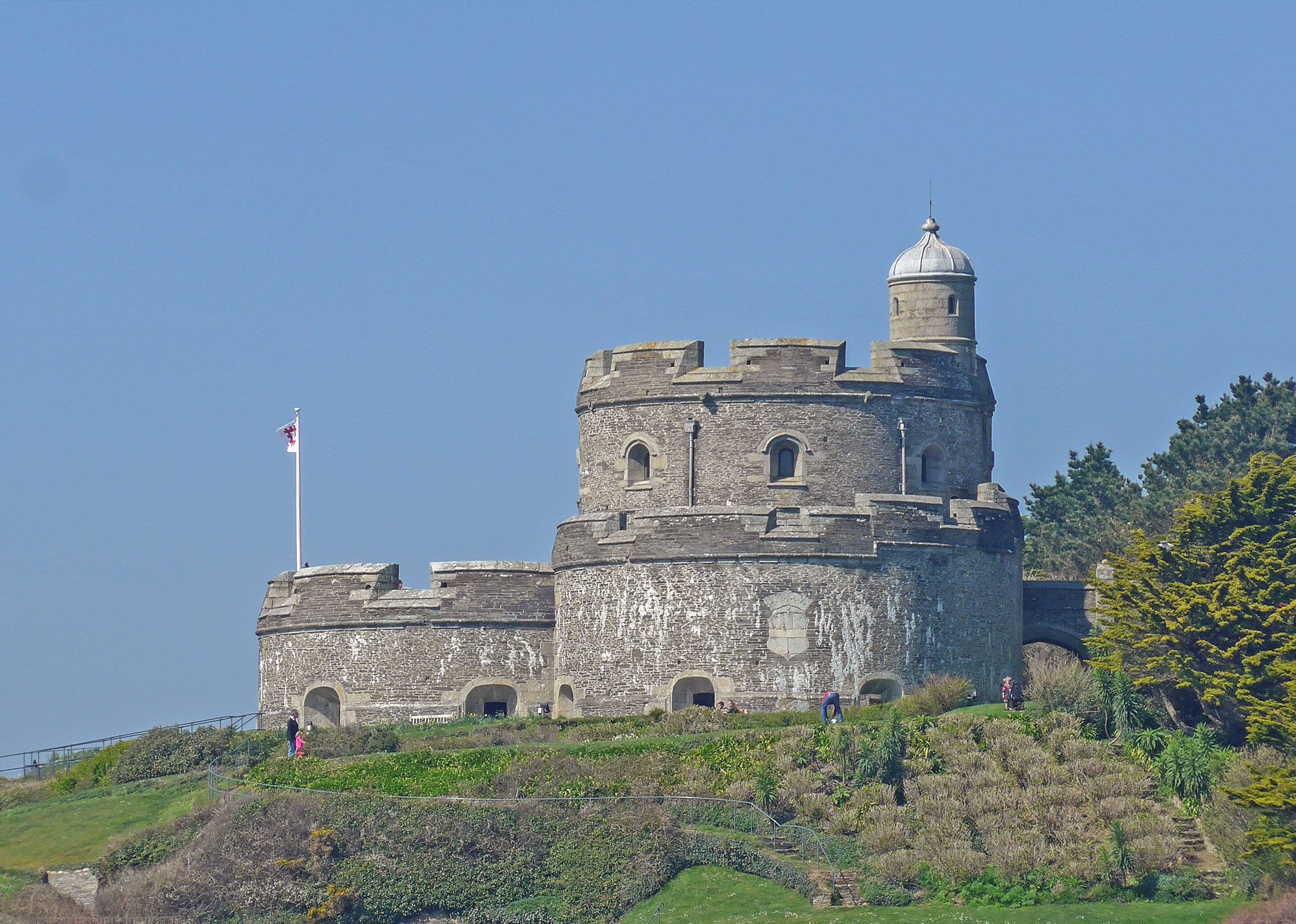 A historic stone castle with a flag on top, surrounded by greenery and hills.