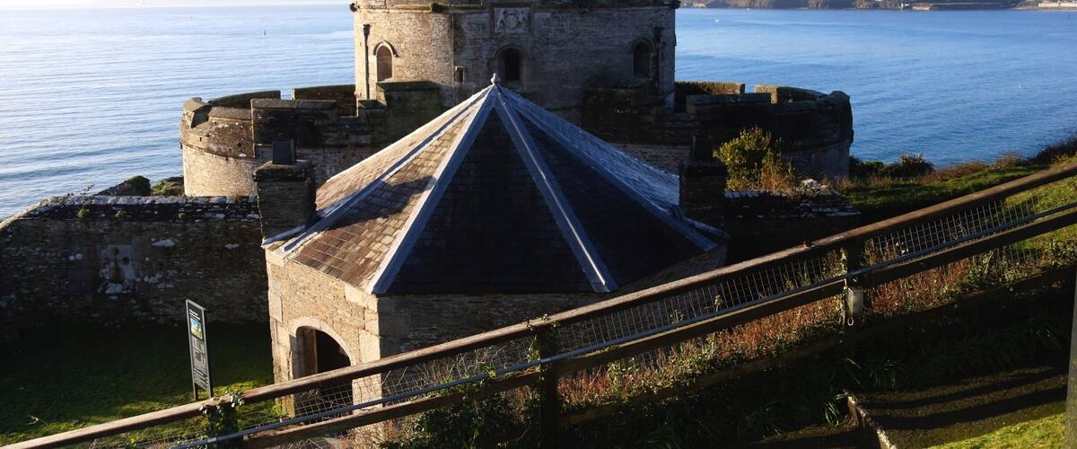 A historic stone fortress with a central tower and a smaller building with a pointed roof, overlooking a calm sea.
