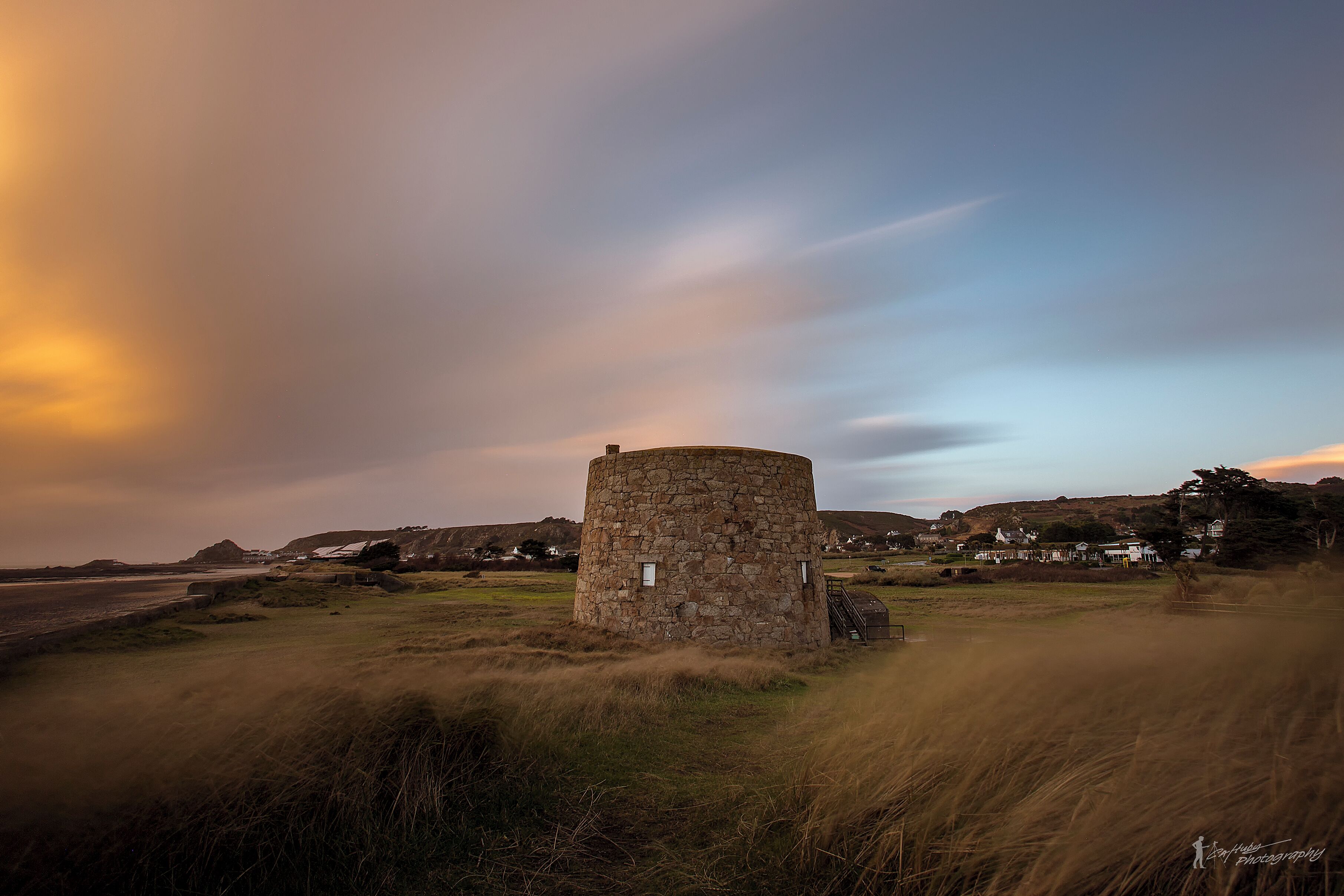 A stone tower in a grassy field with houses in the background.