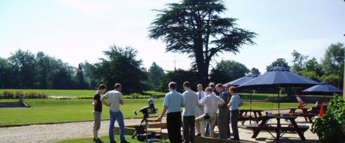 A group of people gathered around a camera on a golf course with a picnic area and a large tree in the background.