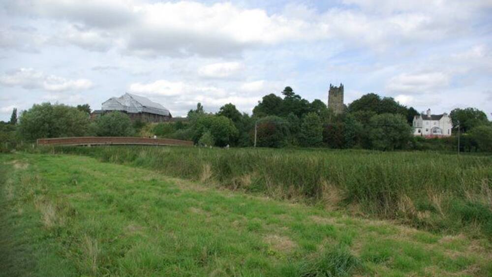 Ein grasbewachsenes Feld mit einer Brücke, einem schlossartigen Bauwerk und einem großen, weißen Gebäude im Hintergrund.